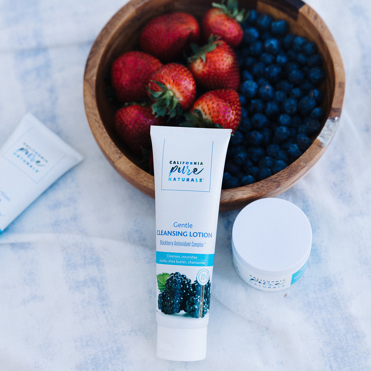Cleansing Lotion and Nourishing Cream products by California Pure Naturals with strawberries and blueberries in a bowl behind products, highlighting the ingredients from which products are made.   